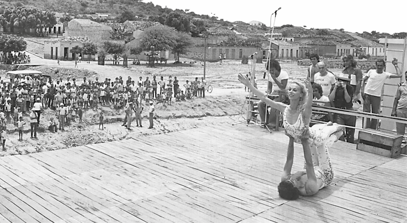 Fotografia em preto e branco. Dançarinos se apresentam em palco de madeira ao ar livre. À frente, um homem está deitado de costas no tablado e suspende o corpo de uma  mulher com as mãos e as pernas. A mulher tem cabelo claro longo e usa uma roupa clara justa. Seus braços estão projetados para a frente. À esquerda, pessoas observam a cena. À direita, ao lado do palco, um grupo de homens acompanha a apresentação. Há um microfone. Ao fundo, céu, árvores, construções e parte de um morro.