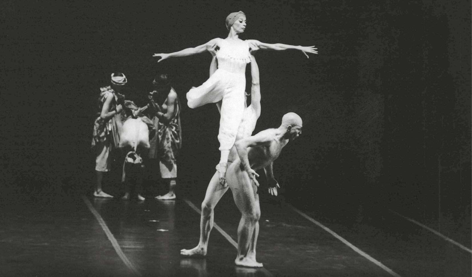 Fotografia em preto e branco. Bailarinos executam movimento de dança em um palco de chão e paredes escuros. Em primeiro plano, uma mulher de turbante, collant e calça brancos. Ela está de braços abertos e é erguida no ar por um homem. À frente deles, um homem está em movimento de dança. Ao fundo, três dançarinos.