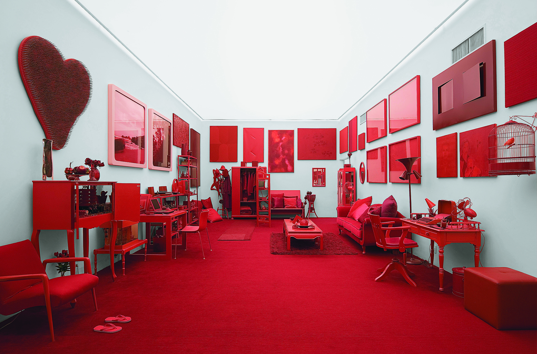 Fotografia. Instalação feita com diversos objetos na cor vermelha em uma sala ampla retangular com paredes cinza claro e teto branco iluminado. Há quadro vermelhos nas paredes e o chão também é vermelho. Há móveis na mesma cor, como sofás, cadeiras, armários, mesas etc.