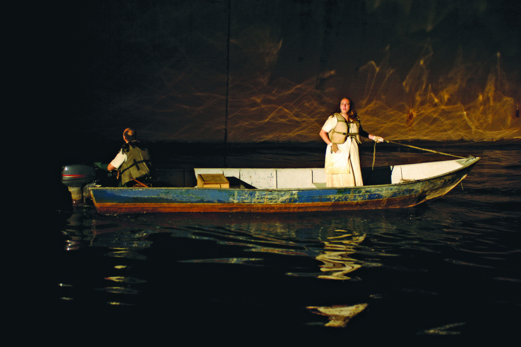 Fotografia. Um barco navegando em um rio com uma mulher de cabelos longos com um vestido branco, em pé, e um homem sentado de costas, responsável por guiar o barco.