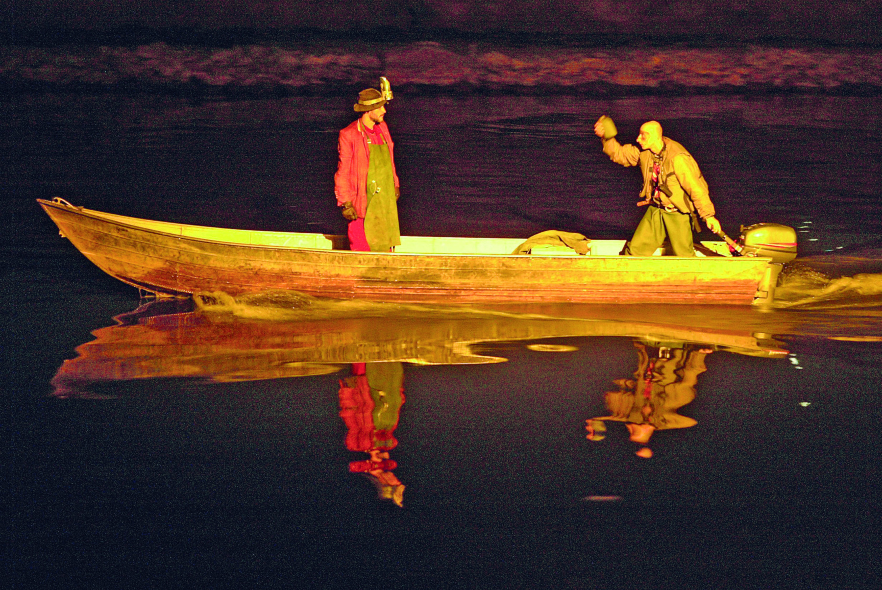 Fotografia. Barco navegando em um rio com um homem de chapéu marrom, vestido com uma camisa e calça vermelhas com um avental marrom por cima, em pé. Manejando o motor do barco, um homem calvo, vestido com casaco e calça marrons, também em pé, gesticula com um dos braços levantados. Suas imagens estão refletidas na água do rio.