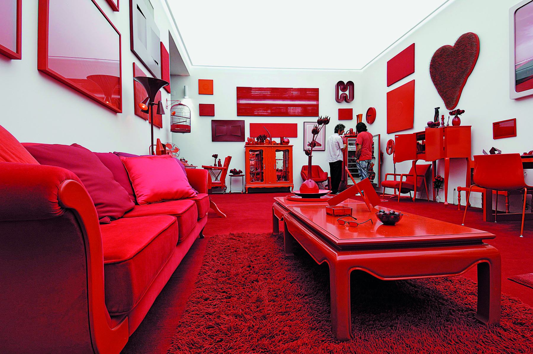 Fotografia. Instalação feita com diversos objetos na cor vermelha em uma sala de paredes cinza claro com teto branco iluminado. Ao fundo, há duas pessoas observando objetos da instalação.