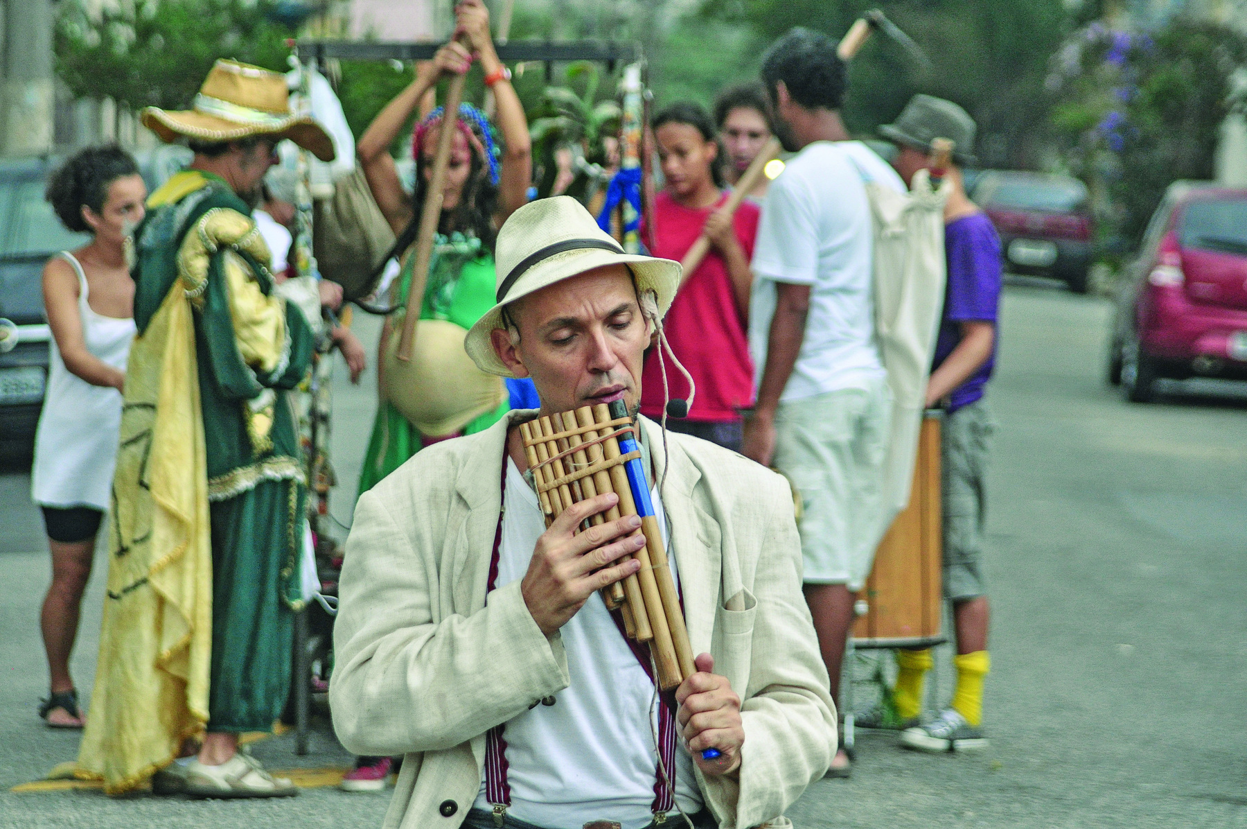 Fotografia. Grupo de atores na rua. Em primeiro plano, um homem de chapéu branco com fita preta, vestido com camiseta e casaco brancos, toca uma flauta de bambu com pequenos cilindros de madeira. Ao fundo, outros atores carregam outros instrumentos musicais.