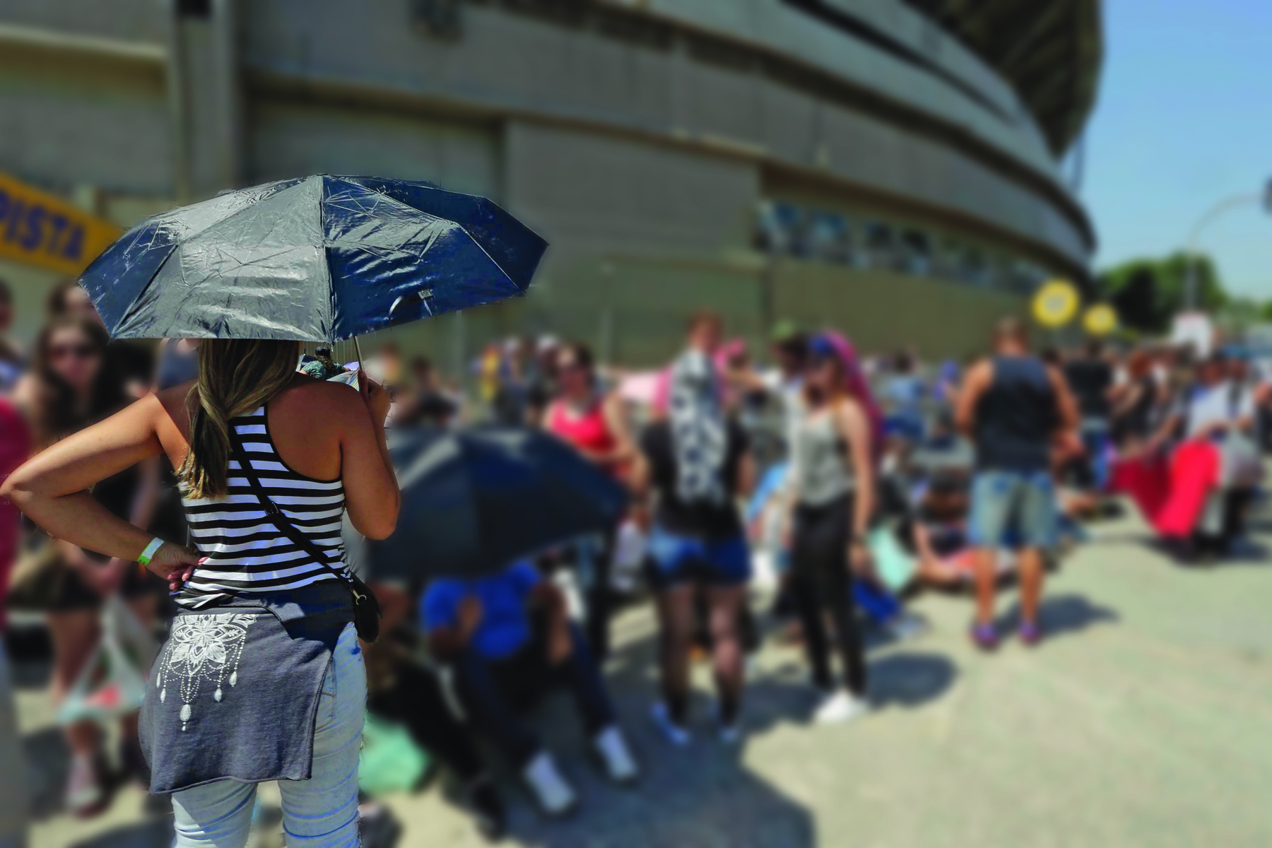 Fotografia. Mulher de guarda-chuva na rua. Ela está de costas e veste camiseta branca com listras horizontais pretas e calça jeans. Está com uma blusa amarrada na cintura e segura um guarda-chuva. Ao fundo, há uma fila de pessoas.