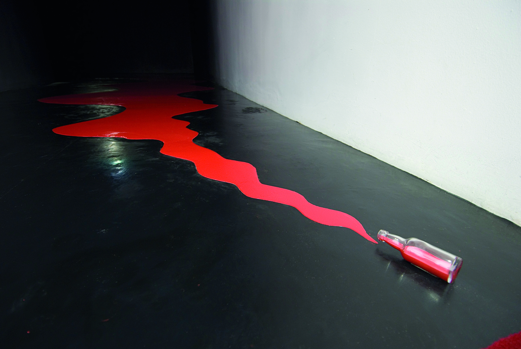 Fotografia. Instalação que apresenta uma garrafa caída que deixa escorrer tinta vermelha em um grande rastro sobre o chão preto.