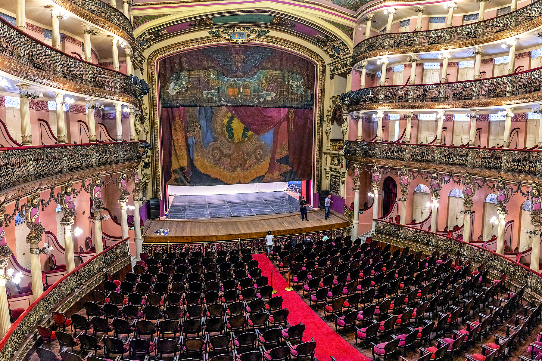 Fotografia. Vista geral da parte interna de um teatro luxuoso, decorado em vermelho e dourado. Há poltronas na plateia e quatro andares de camarotes nas laterais. O palco é grande e tem uma cortina em tons de vermelho e amarelo.