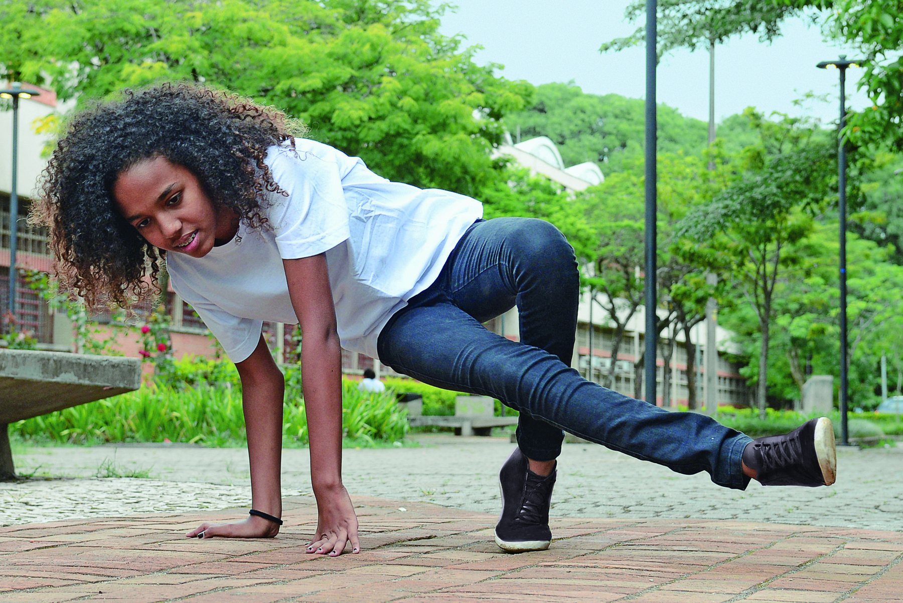 Fotografia. Menina de cabelo cacheado castanho, vestida com camiseta branca e calça azul, executa um movimento rente ao solo, com os dois braços e uma perna apoiados e a outra perna esticada, paralela ao chão.