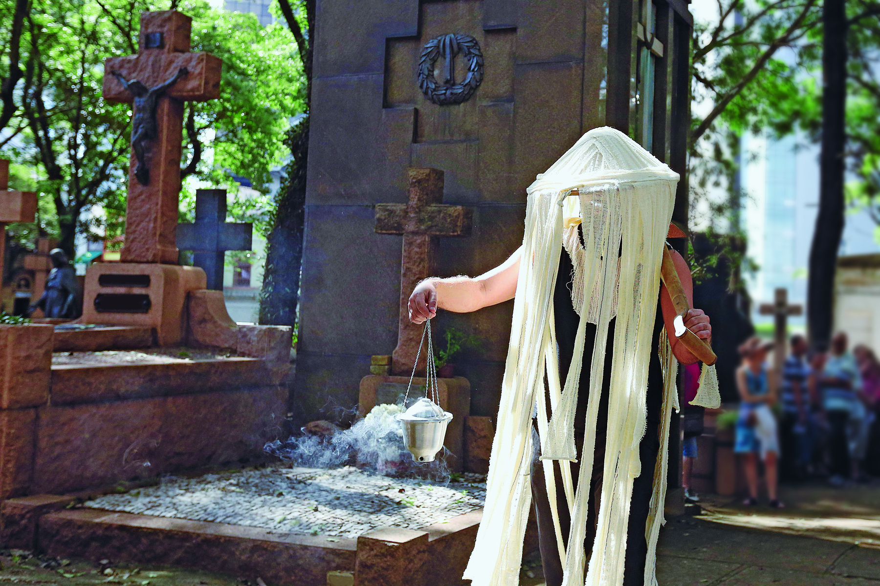 Fotografia. Atriz representando uma peça em frente a um túmulo, vestida com um chapéu de tecido branco que cobre seu rosto e parte do corpo. Ela segura em uma das mãos um incensário, que solta uma fumaça branca. Ao fundo, algumas pessoas observam a cena.