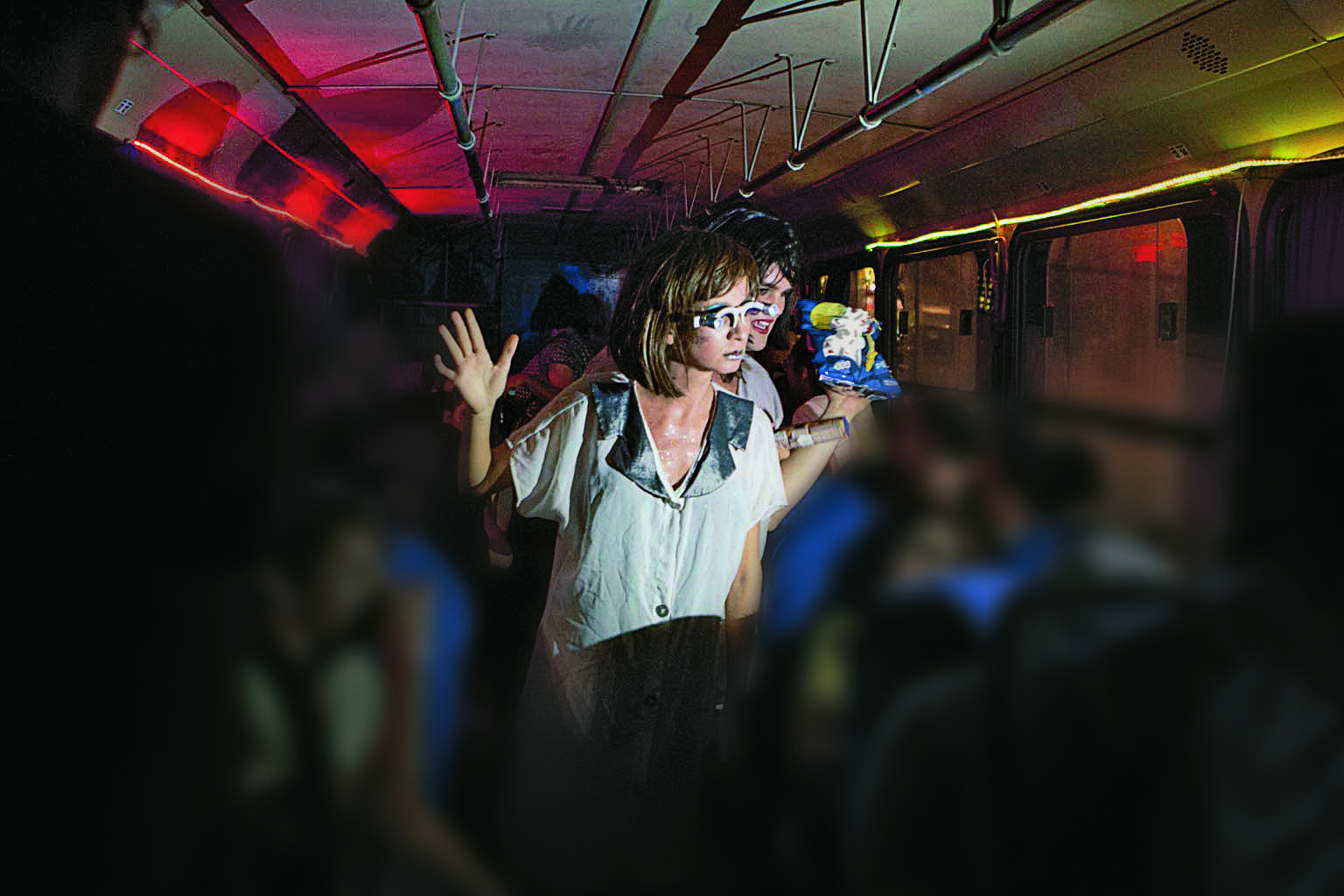 Fotografia. Atrizes interpretam uma cena dentro de um ônibus. Destaque para uma mulher de cabelo loiro e óculos que caminha no corredor do coletivo com objetos nas mãos.