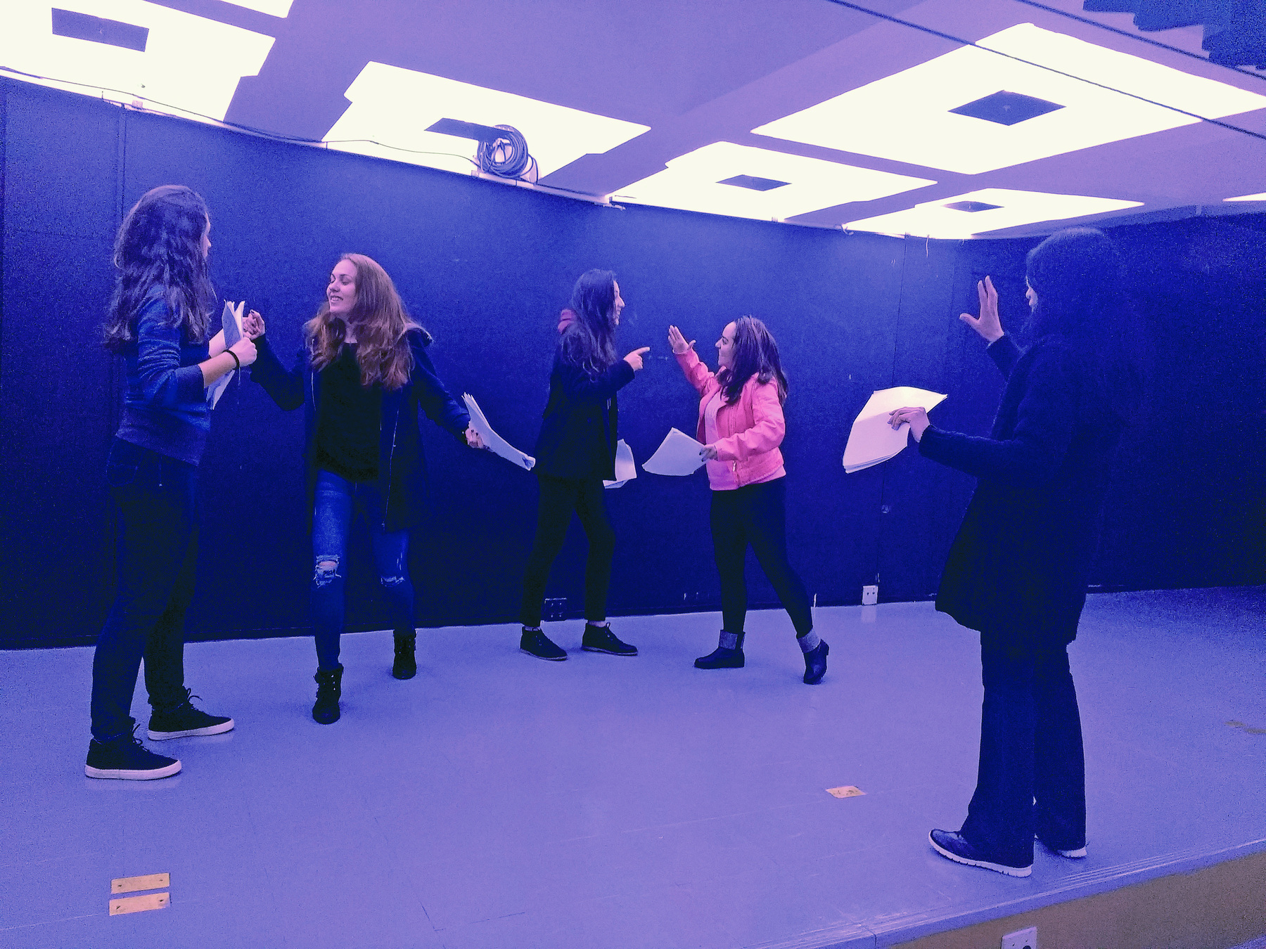 Fotografia. Quatro atrizes em uma sala escura, de paredes pretas, ensaiam em duplas com papéis nas mãos, enquanto são observadas por uma mulher que faz um gesto com as mãos.