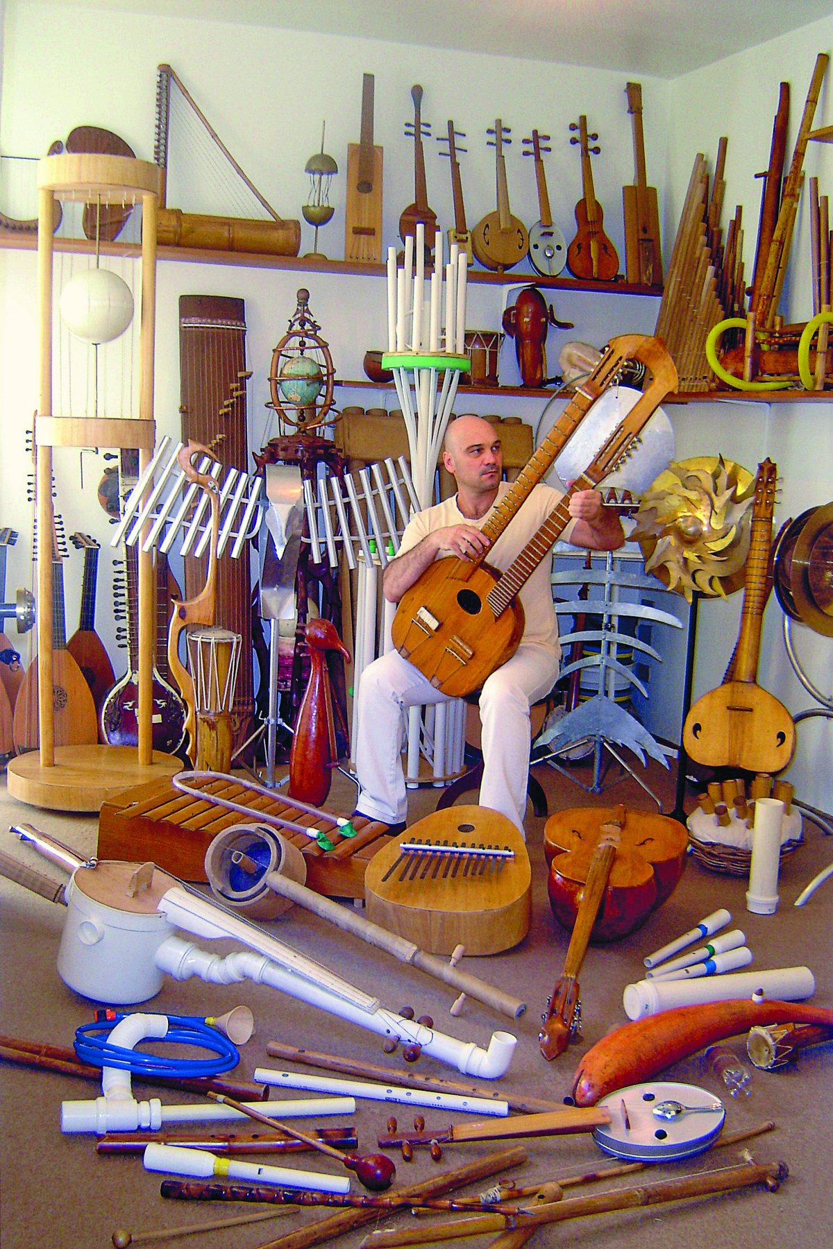 Fotografia. Homem calvo, vestindo camiseta amarela e calça branca, sentado em um banco. Ao seu redor, há diversos instrumentos musicais diferentes, como instrumentos de sopro feitos com tubos de PVC.