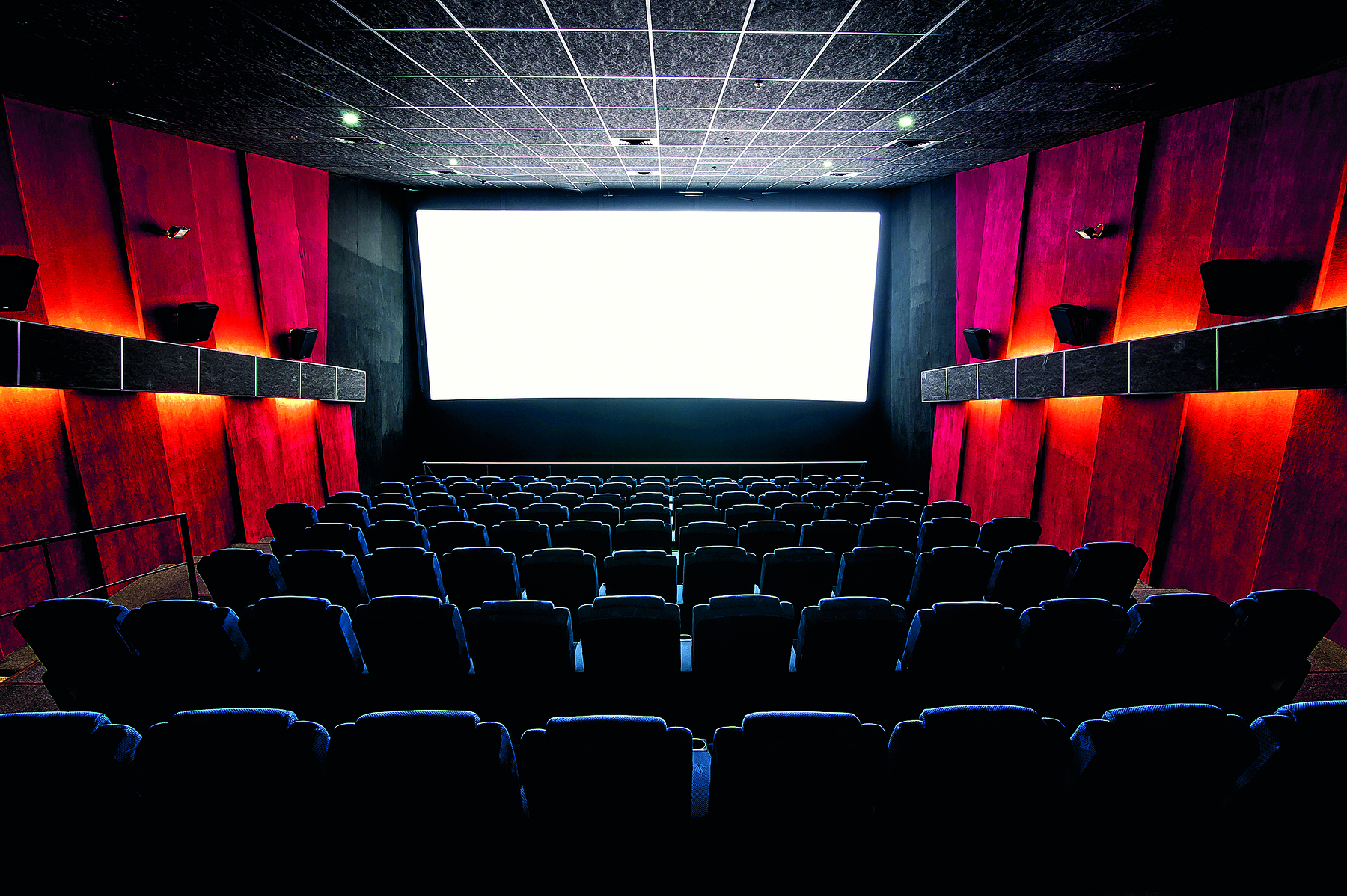 Fotografia. Sala de cinema vazia. Em primeiro plano, poltronas da plateia. Ao fundo, no centro, a tela de projeção. Nas laterais da sala, há caixas de som.