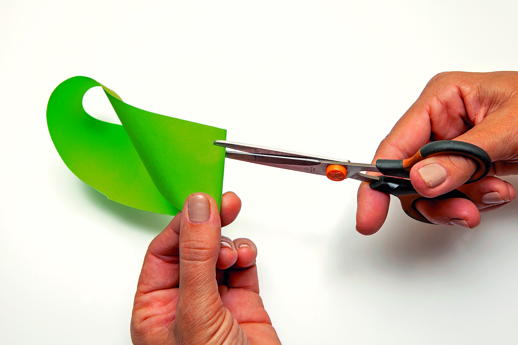 Fotografia. Detalhe de duas mãos cortando parte da fita de papel verde retorcida.