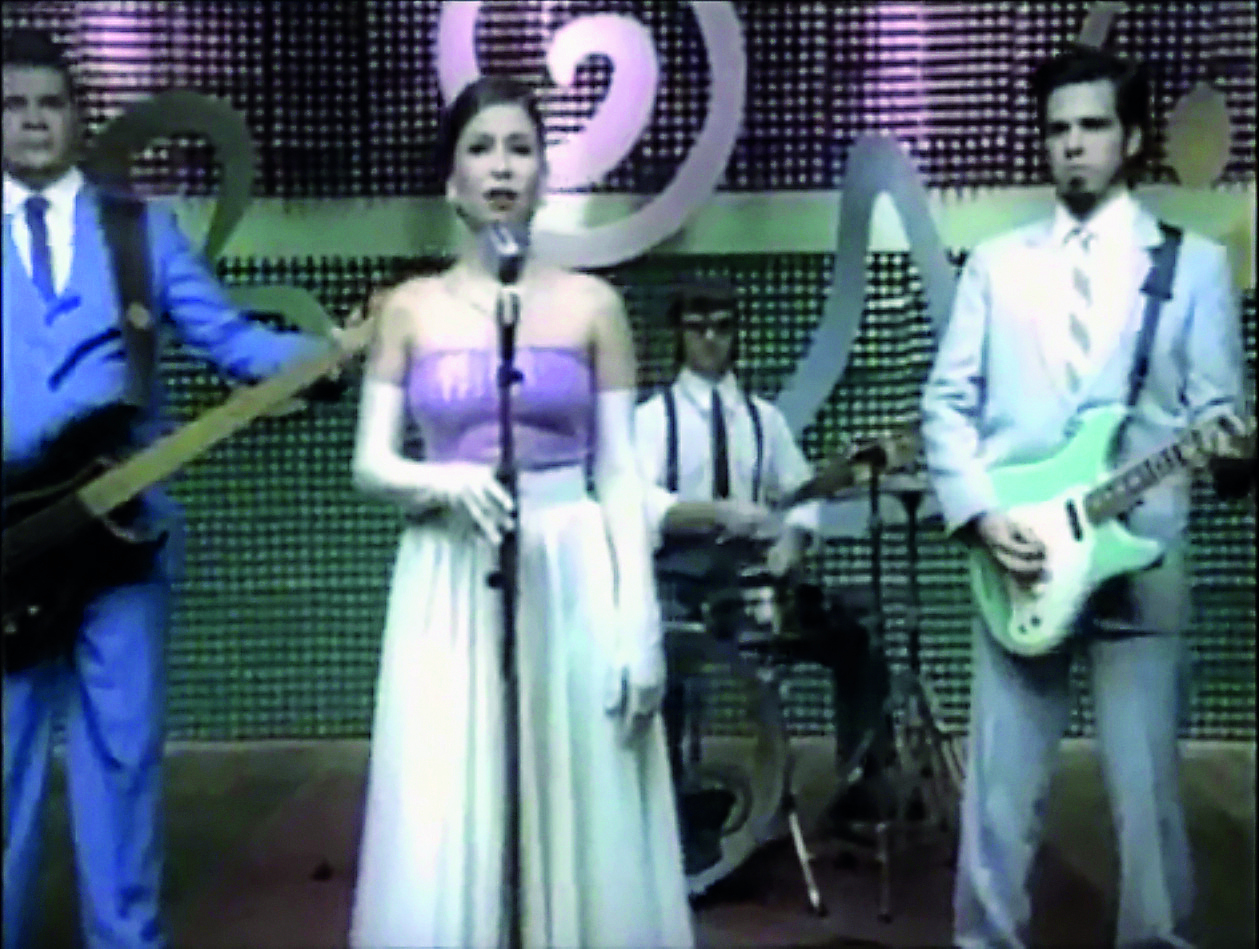 Fotografia. Show musical com uma cantora e uma banda de apoio composta de três músicos. A cantora está usando um vestido clássico nas cores rosa e branco e usa luvas brancas que vão acima do cotovelo. Os músicos, ao fundo, vestem terno e gravata.
