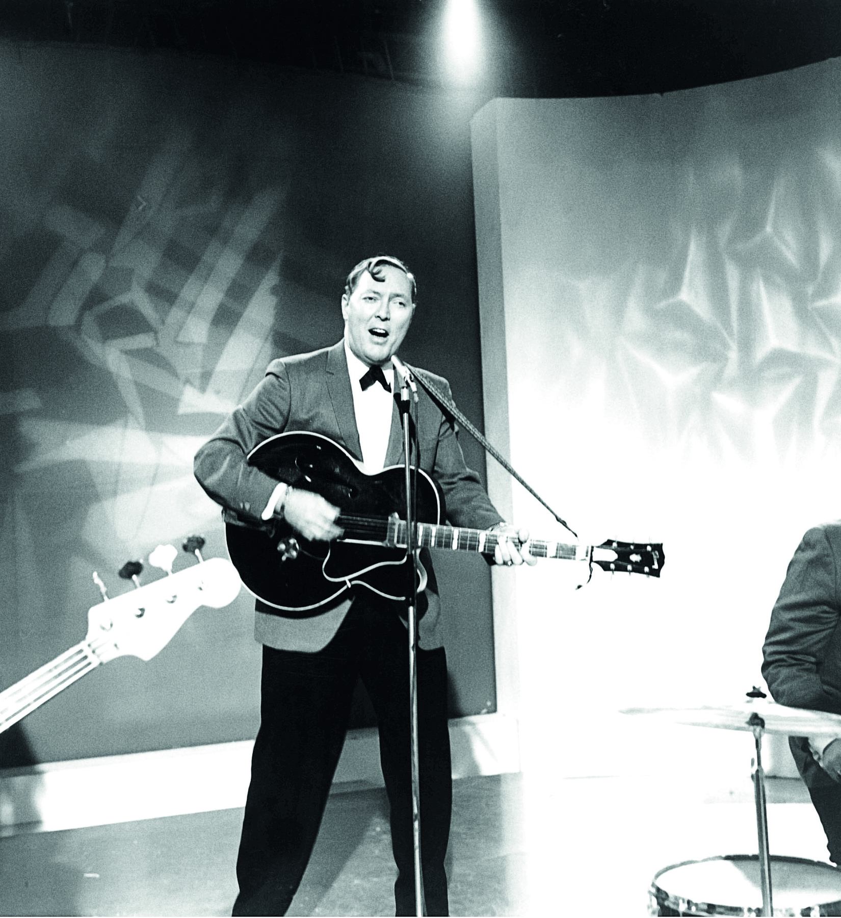 Fotografia em preto e branco. Homem de cabelo curto, vestindo camisa, gravata borboleta e paletó, canta e toca violão com um microfone à frente.