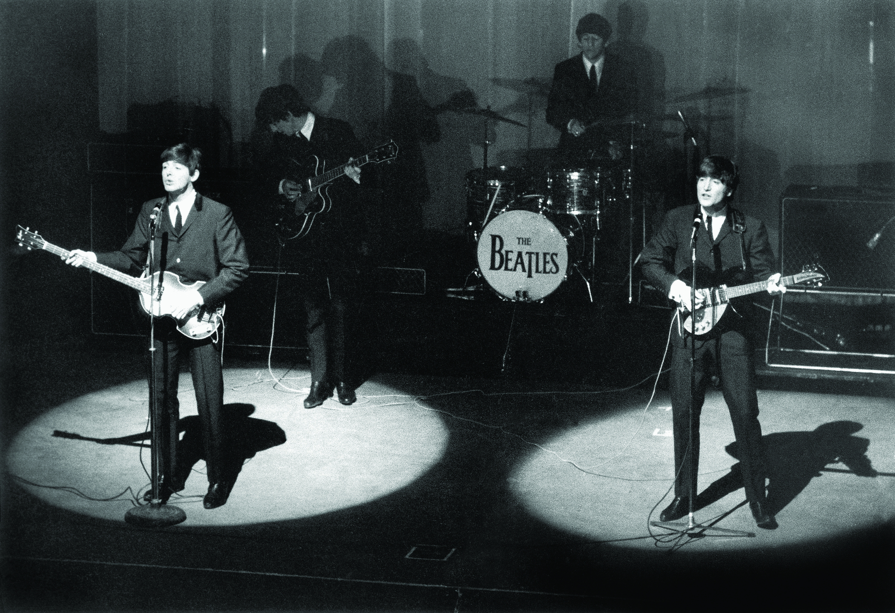 Fotografia em preto e branco. Show musical com quatro músicos no palco, todos usam cabelos curtos e vestem paletó e gravata. Os dois músicos em primeiro plano cantam e tocam. Ao fundo, no bumbo da bateria, há o logotipo da banda: “The Beatles”.