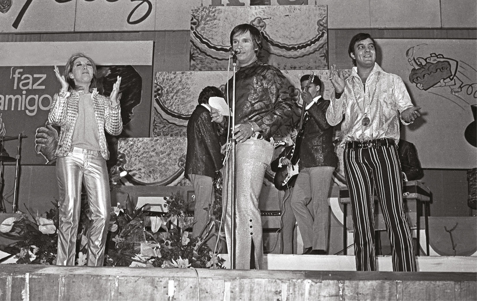 Fotografia em preto e branco. Músicos em um palco, cantando e batendo palmas. À esquerda, uma mulher de cabelo preso, vestida com casaco e calça brilhantes, está batendo palma. No centro, um homem vestido com uma camisa e calça, canta ao microfone. À direita, um homem de cabelo curto, vestido com camisa brilhante e calça com listras verticais, também bate palmas. Ao fundo, há dois músicos vestidos com terno.