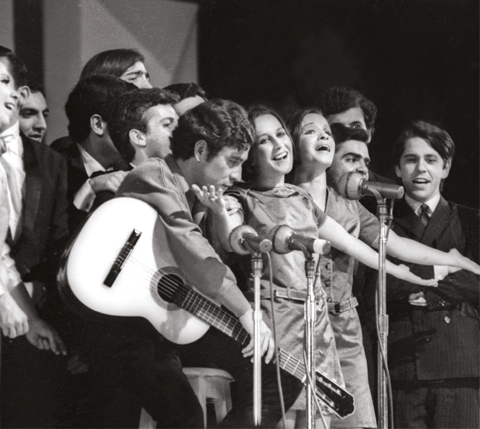 Fotografia em preto e branco. Homens e mulheres cantam e tocam juntos em um palco. Há homens e mulheres, que dividem três microfones. Destaque para um dos músicos que segura um violão e tem a cabeça baixa.