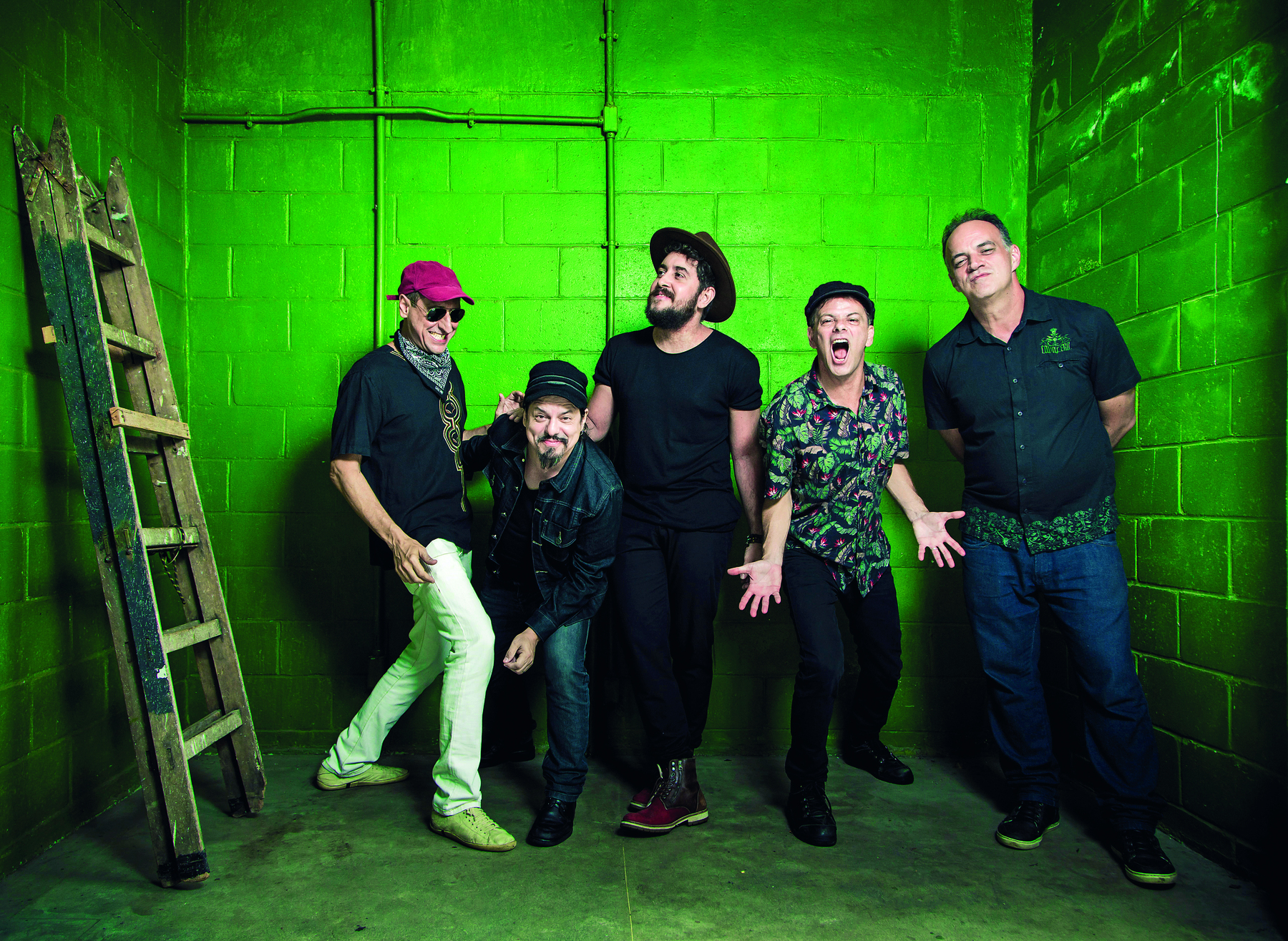 Fotografia. Cinco homens em uma sala de paredes verdes. Eles fazem pose para a foto. Quatro deles usam bonés ou chapéu. À esquerda, há uma escada de madeira apoiada na parede.