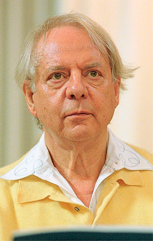 Fotografia. Homem de cabelo grisalho, vestido com camisa branca com um casaco amarelo.