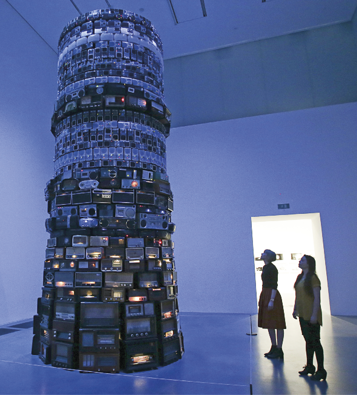 Fotografia. Duas mulheres observam uma instalação composta de uma grande torre construída com diferentes tipos de rádio, antigos e novos, no interior de um salão com paredes altas.