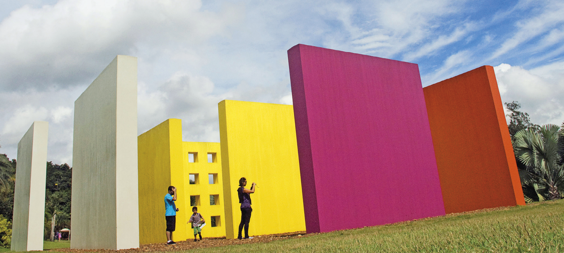 Fotografia. Instalação a céu aberto. Há grandes paredes nas cores rosa, laranja, amarelo e branco no meio do parque. Entre as paredes, há pessoas que observam a obra e tiram fotos. Ao fundo, uma parede diferente na cor amarela com parede que apresenta orifícios quadrados.