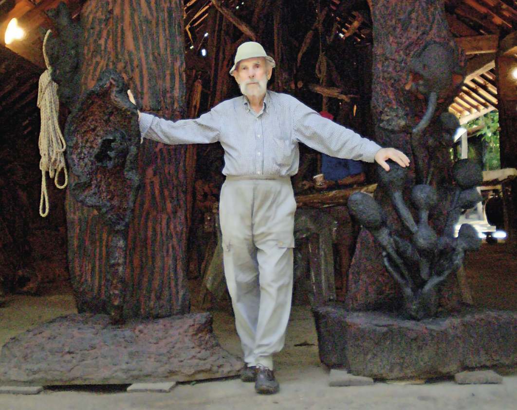 Fotografia. Homem de barba grisalha, vestido com camisa azul-claro e calça cinza, além de um chapéu também na cor azul-claro, posa ao lado de esculturas abstratas de madeira escura.