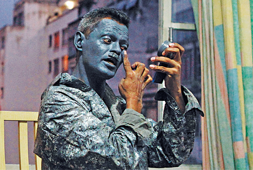 Fotografia. Homem de cabelo curto pinta o rosto com tinta azul, e está vestido com roupas da mesma cor da tinta, se preparando para fazer estátua viva.