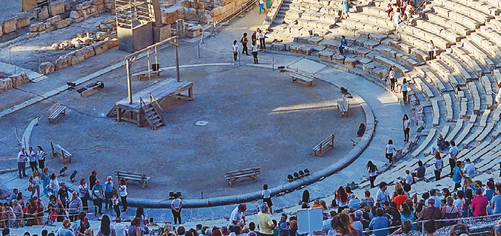 Fotografia. Vista superior de um teatro a céu aberto, feito de pedra, com destaque para o palco. Nele, há um palco menor, feito de madeira.