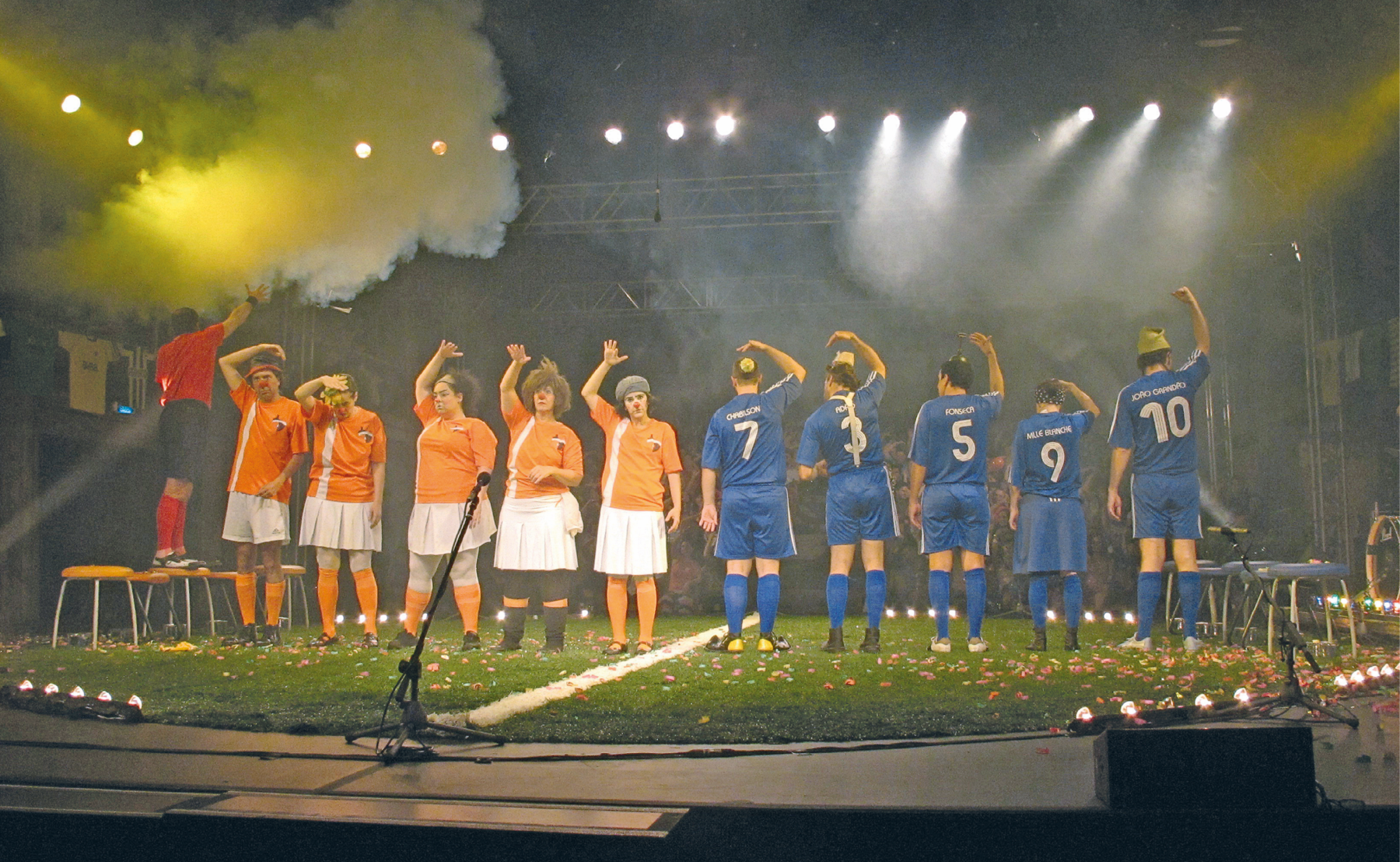 Fotografia. Palco com formato de campo de futebol pequeno, com gramado e jogadores de dois times: um com uniforme composto por camiseta laranja e saia branca e o outro com uniforme todo azul. Há um juiz em cima de uma cadeira com uniforme vermelho e preto. Os jogadores parecem cumprimentar o público do estádio. Há um jogo de luz e fumaça no ambiente.