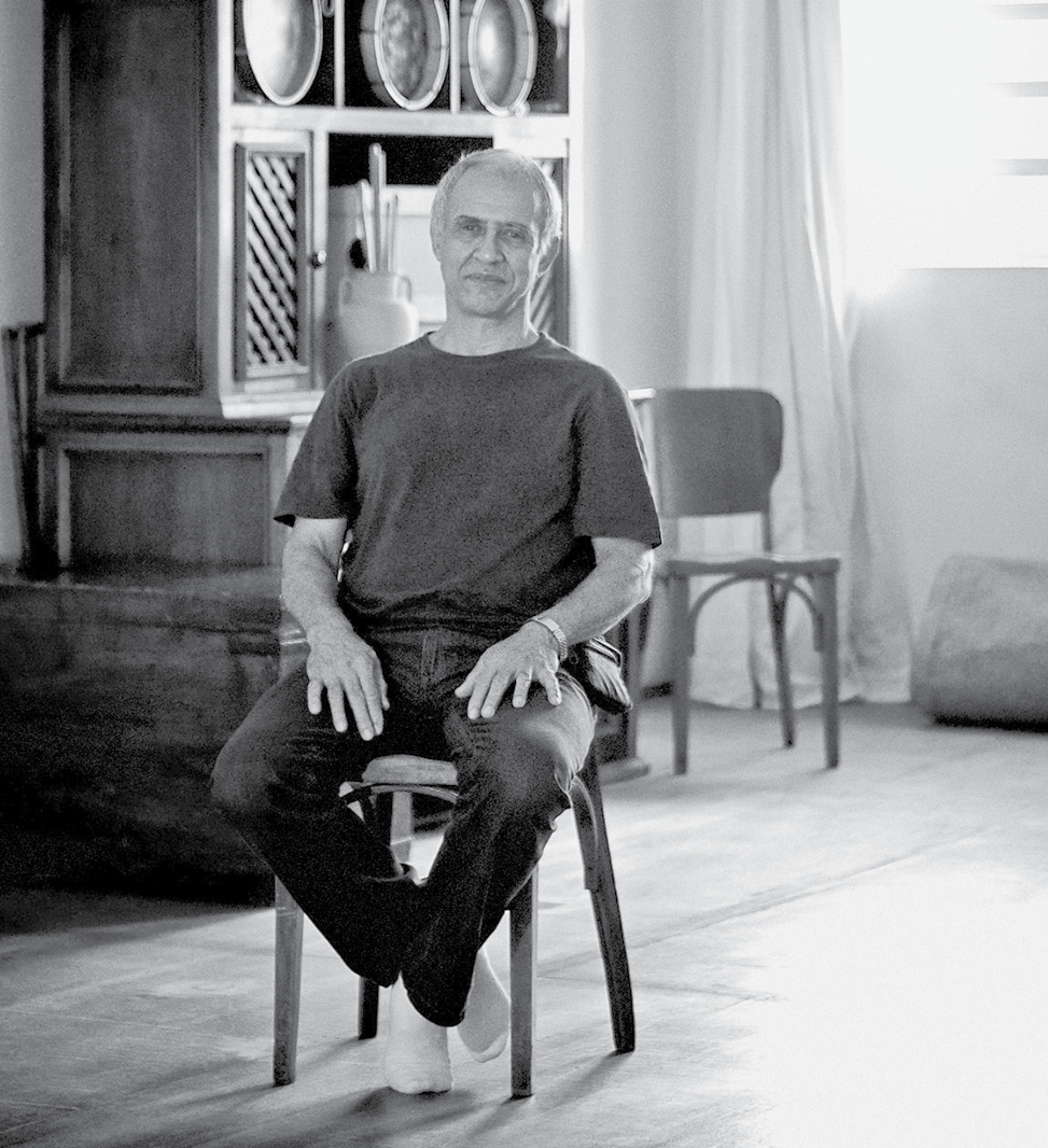 Fotografia em preto e branco. Homem de cabelo curto grisalho, vestido com camiseta e calça escuras. Está sentado em uma cadeira no centro de uma sala. Ao fundo, uma estante e uma cadeira de madeira.