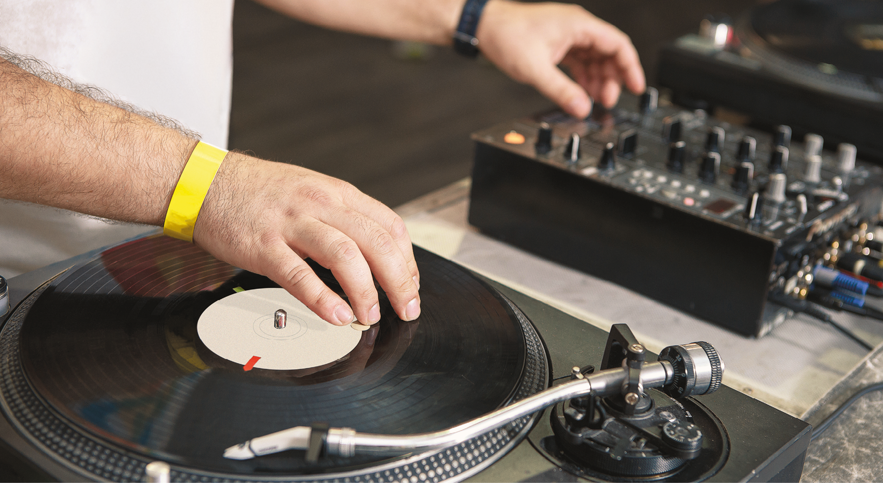 Fotografia. Detalhe das mãos de um DJ, que ajusta o volume em uma mesa de som ao mesmo tempo em que movimenta um LP ou vinil no toca-discos.