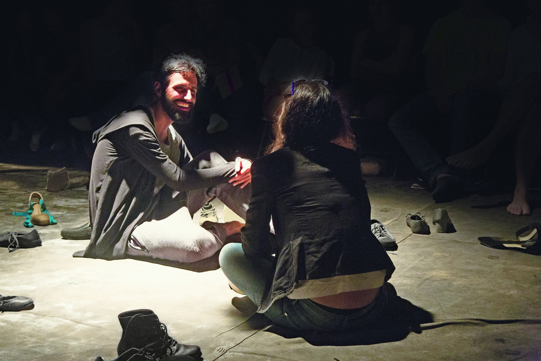 Fotografia. Duas pessoas conversam sentadas ao chão. Um homem de cabelo e barba castanhos, vestido com uma blusa cinza e uma mulher, de costas, vestida com um casaco preto e calça jeans, rodeados por sapatos espalhados no chão.