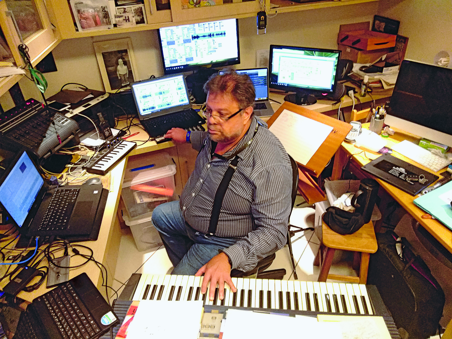 Fotografia. Homem de óculos com cabelo curto castanho, vestido com uma camisa com tons de cinza. Ele está sentado entre um teclado e telas de computadores, entre outros aparelhos eletrônicos.