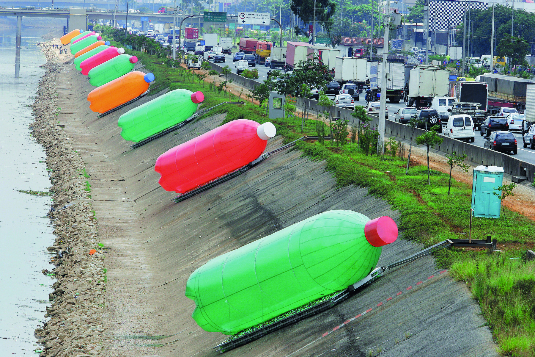 Fotografia. Intervenção artística localizada à beira de um rio de cidade grande com esculturas gigantes coloridas com o formato de garrafas pet. Ao lado, trânsito intenso de carros em uma via expressa.