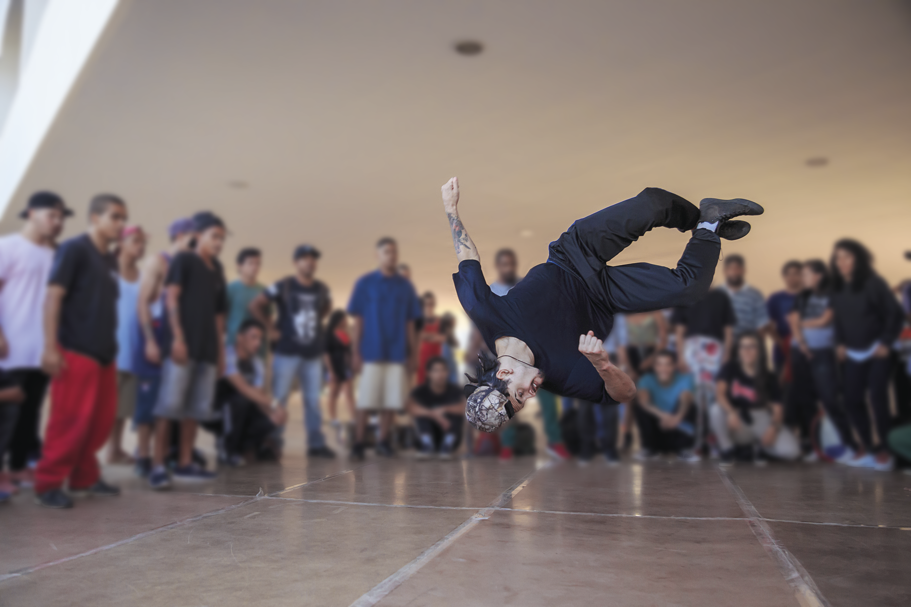 Fotografia. Homem executa um movimento de dança. Vestido com roupas pretas, ele está no ar, com a cabeça apontada para o chão, com as pernas cruzadas ao alto e os braços semiabertos em um salão com pessoas observando seus movimentos.