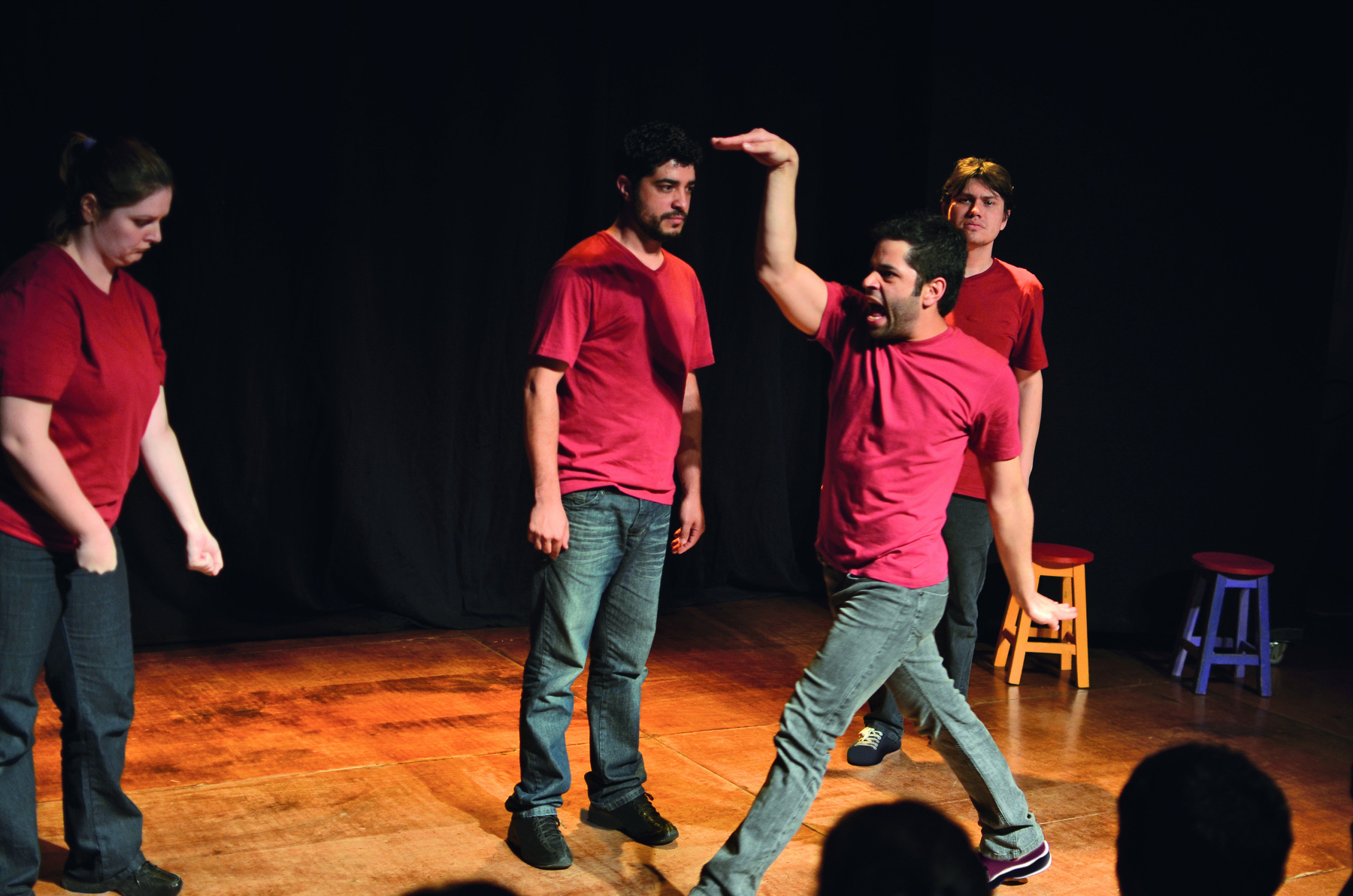 Fotografia. Quatro atores encenam em um pequeno palco. Eles vestem camisetas vermelhas e calças jeans. À esquerda, uma atriz e, à direita, três atores. Um deles, em primeiro plano, está andando e movendo os braços.