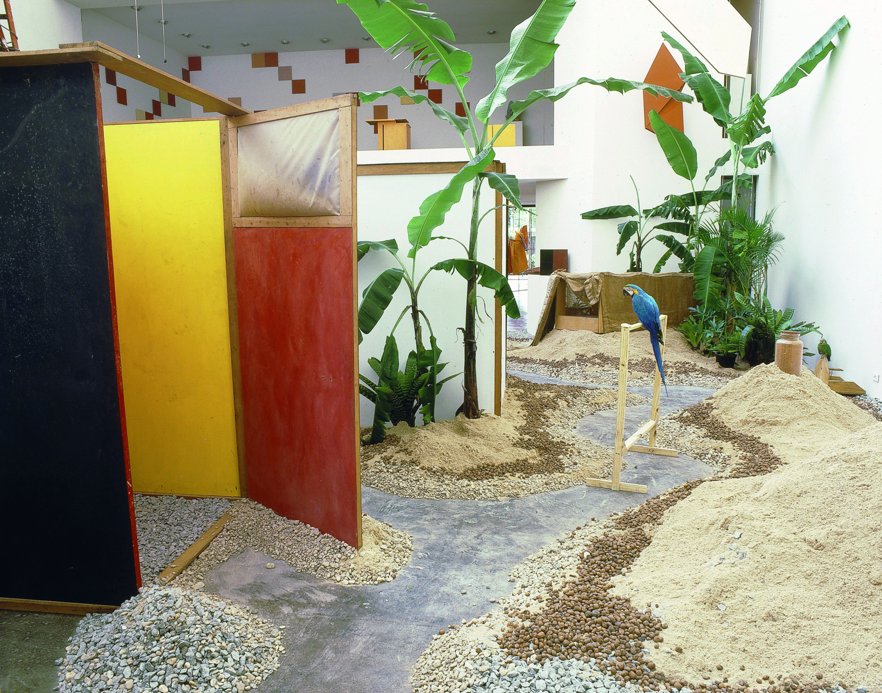 Fotografia. Instalação que apresenta um espaço com areia e pequenos amontoados de pedras no chão, além de tapumes de madeira, que separam o espaço em pequenos ambientes. Há plantas diversas e, em um poleiro, uma arara-canindé.