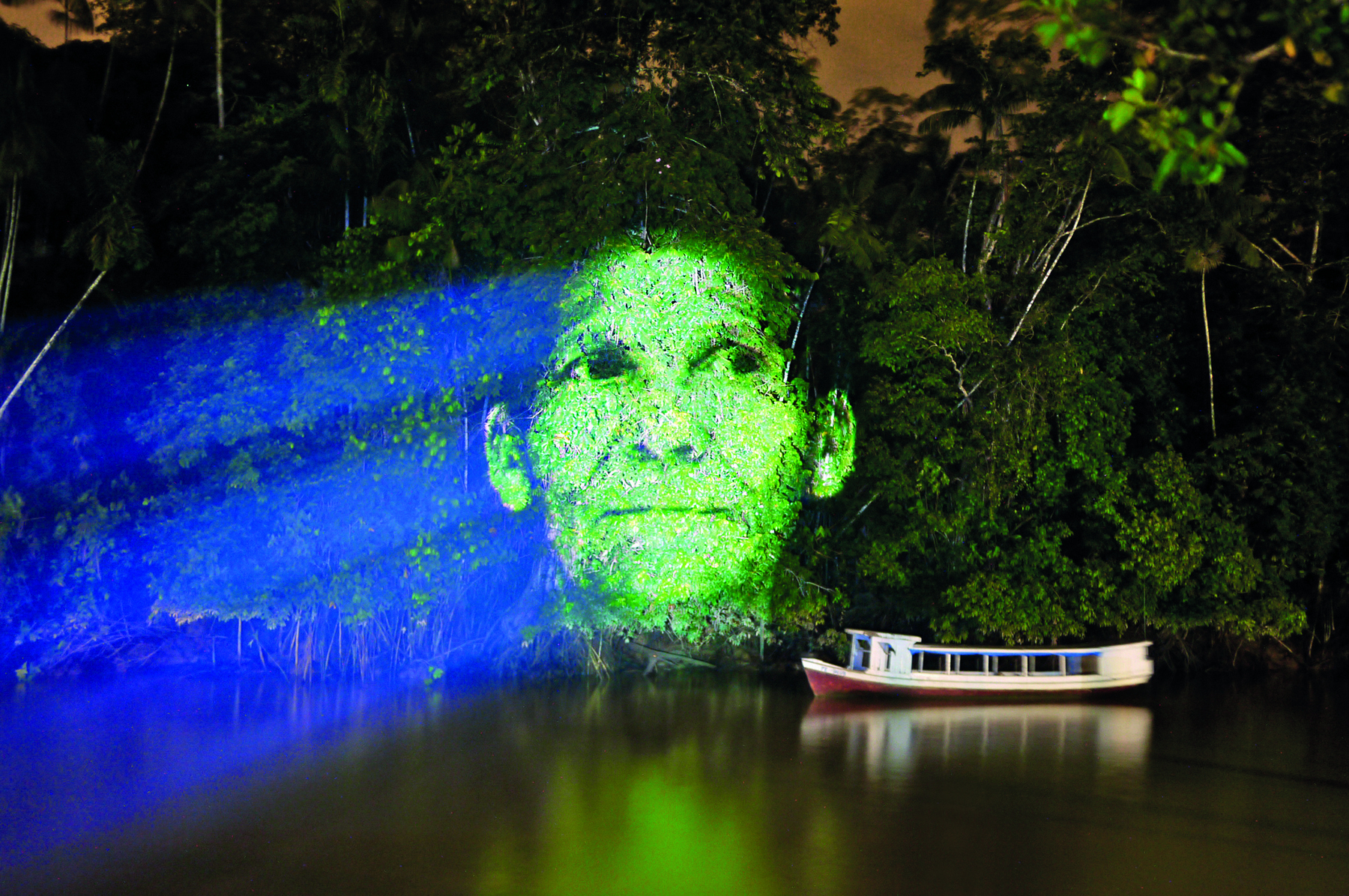 Fotografia. Projeção da imagem do rosto de uma pessoa na mata de um rio, durante o período noturno. Essa projeção tem tons de verde. À direita, um pequeno barco de passageiros navega no rio.