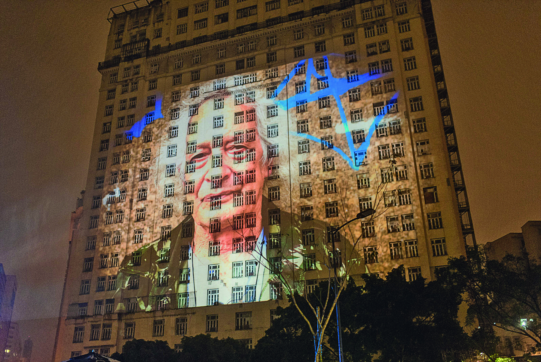 Fotografia. Projeção do rosto de uma pessoa na fachada de um prédio alto durante o período noturno. Trata-se de um homem idoso de cabelo grisalho, vestido com uma camiseta branca e um casaco preto.