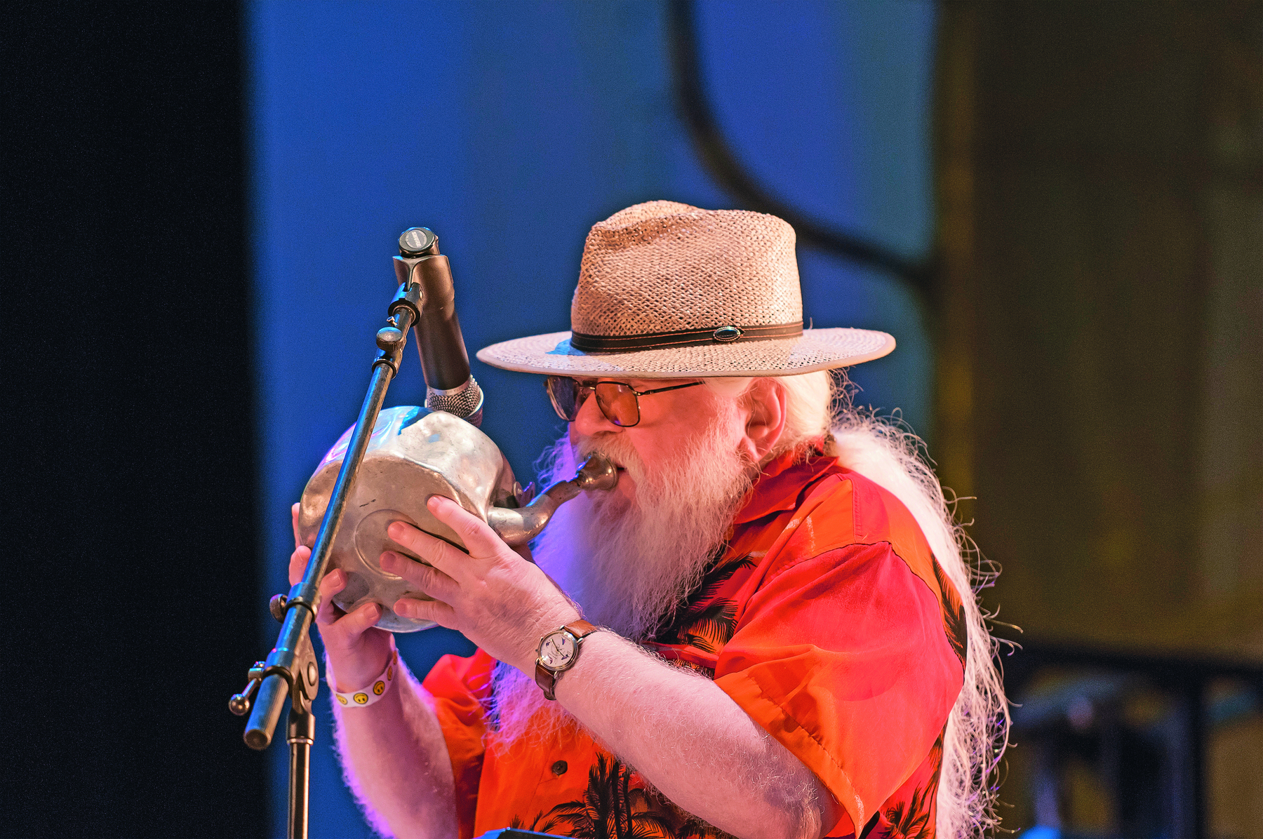 Fotografia. Homem de cabelo e barba longos e grisalhos, com chapéu de palha, vestido com uma camisa laranja, toca um instrumento de metal em formato de chaleira diante de um microfone.