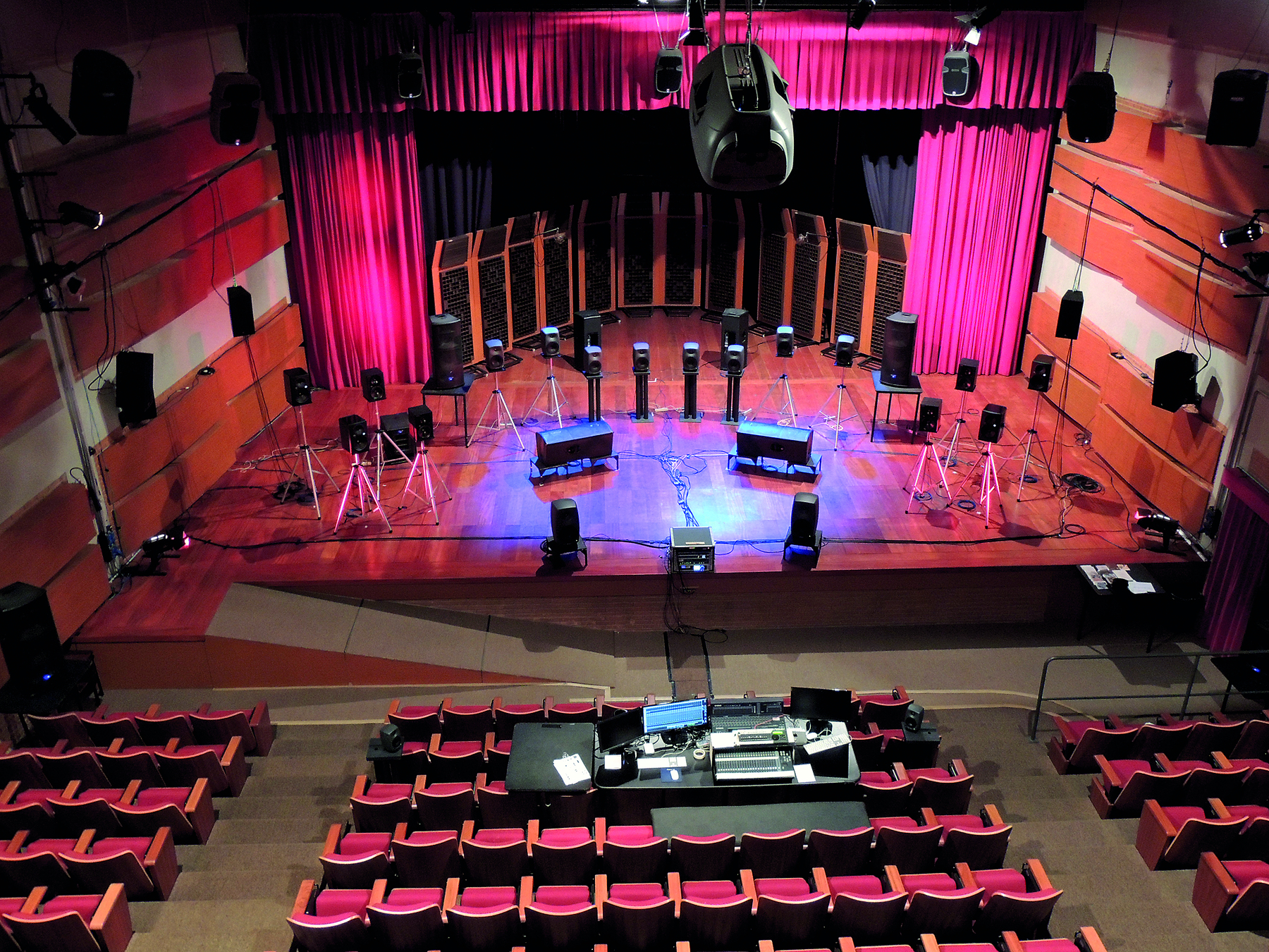 Fotografia. Vista do alto de um palco com alto-falantes em pedestais posicionados como se fosse uma orquestra. A plateia está vazia e, ao fundo, há cortinas vermelhas.