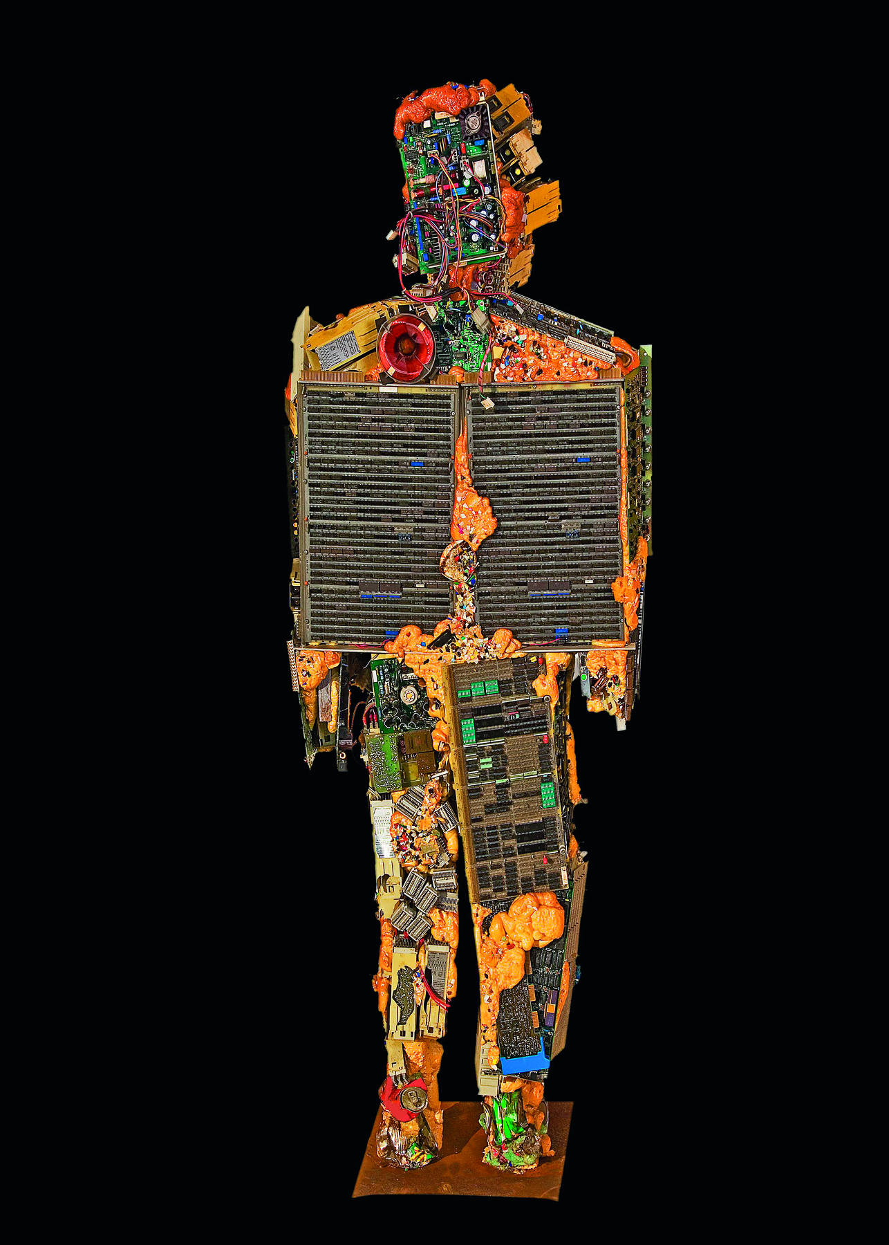 Fotografia. Escultura feita com lixo cujo formato lembra uma pessoa. Há diversos tipos de objetos, entre eles, restos de placas de computador e transistores.