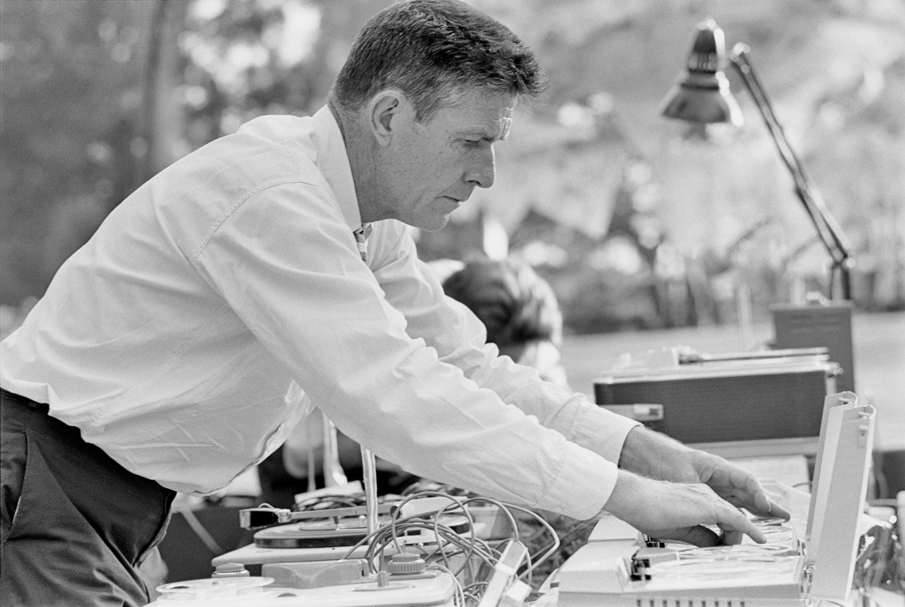 Fotografia em preto e branco. Homem de perfil, vestido com uma camisa branca, mexe em uma mesa de controle de som.