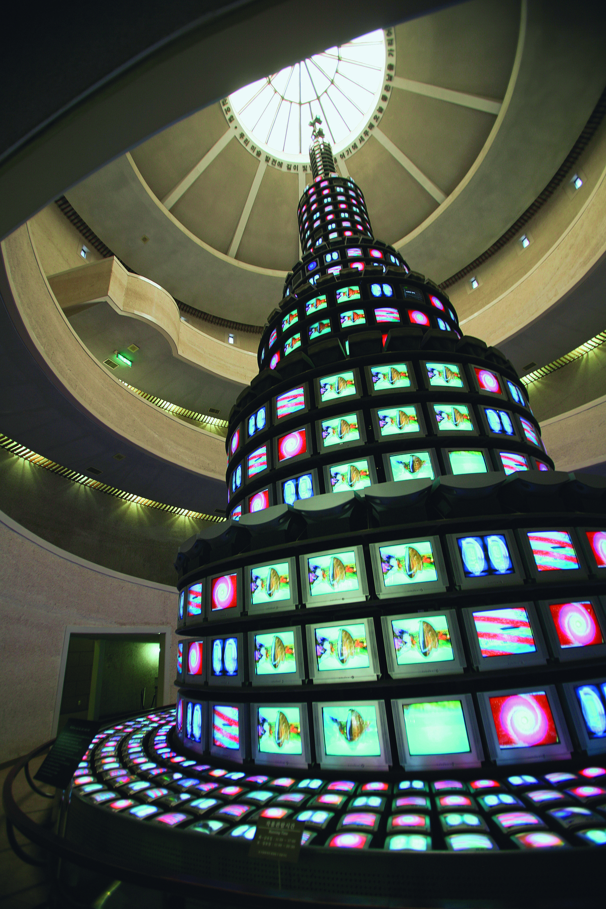 Fotografia. Videoinstalação feita com aparelhos de TV ligados. Trata-se de uma torre alta, com mais de 10 níveis, que quase alcança o teto. Na base da obra, há monitores de TV deitados, formando uma espécie de pedestal para a torre. As cores das imagens mostradas nos monitores de TV são  vermelho e azul-claro. As imagens são abstratas. Ao fundo, salão com mezaninos para que as pessoas possam apreciar as camadas superiores da obra de arte.