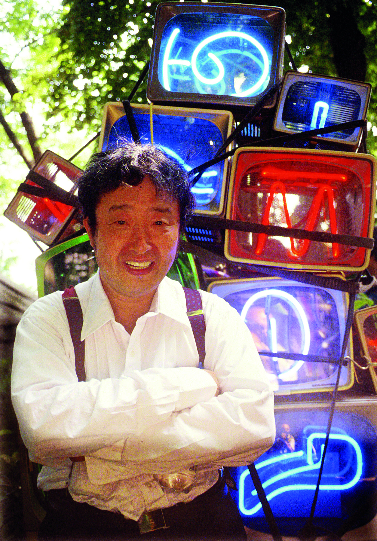 Fotografia. Homem de cabelo curto preto, vestido com camisa branca e suspensórios na cor marrom. Ao fundo, objetos abstratos formados por lâmpadas coloridas de neon.