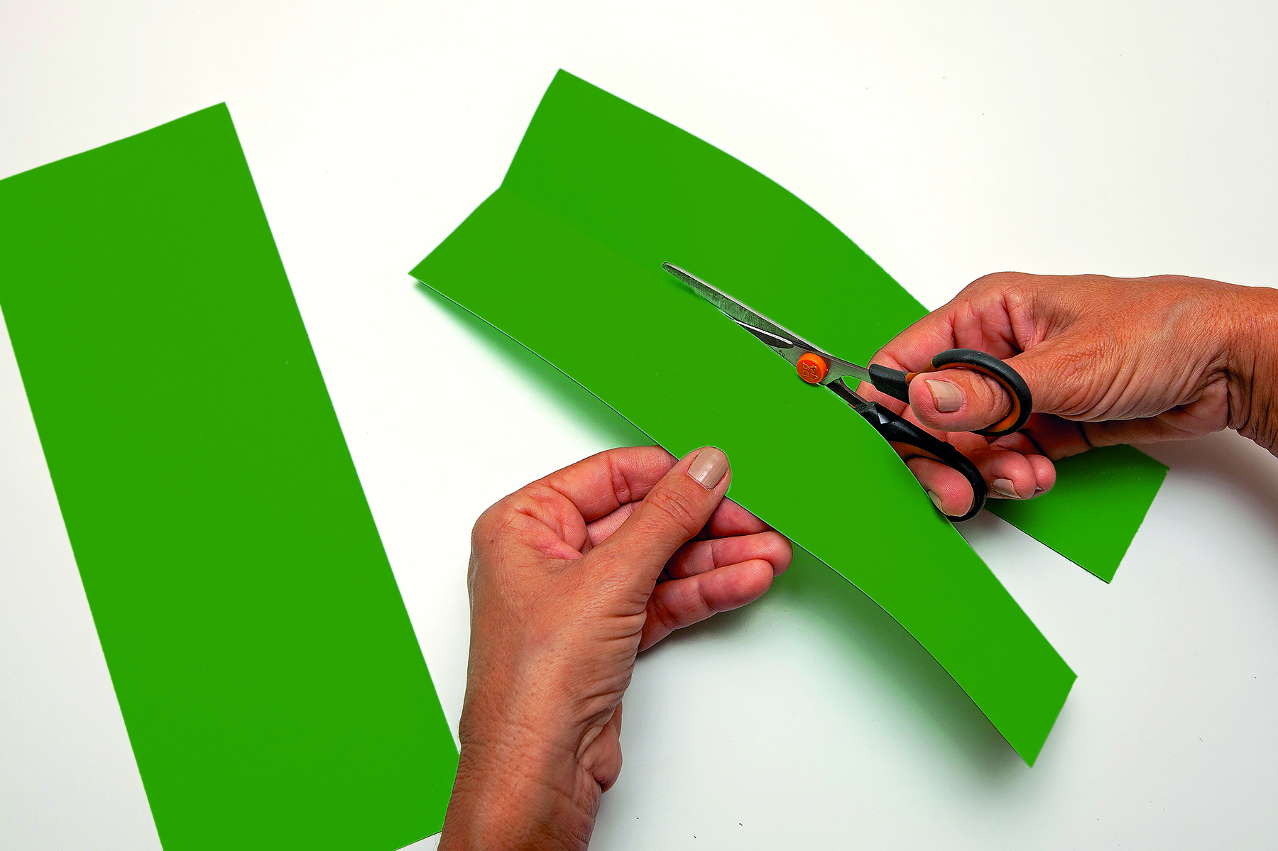 Fotografia. Detalhe de mãos recortando tiras de papel verde com tesoura. O papel contém uma dobradura bem ao centro, que guia o recorte com a tesoura.