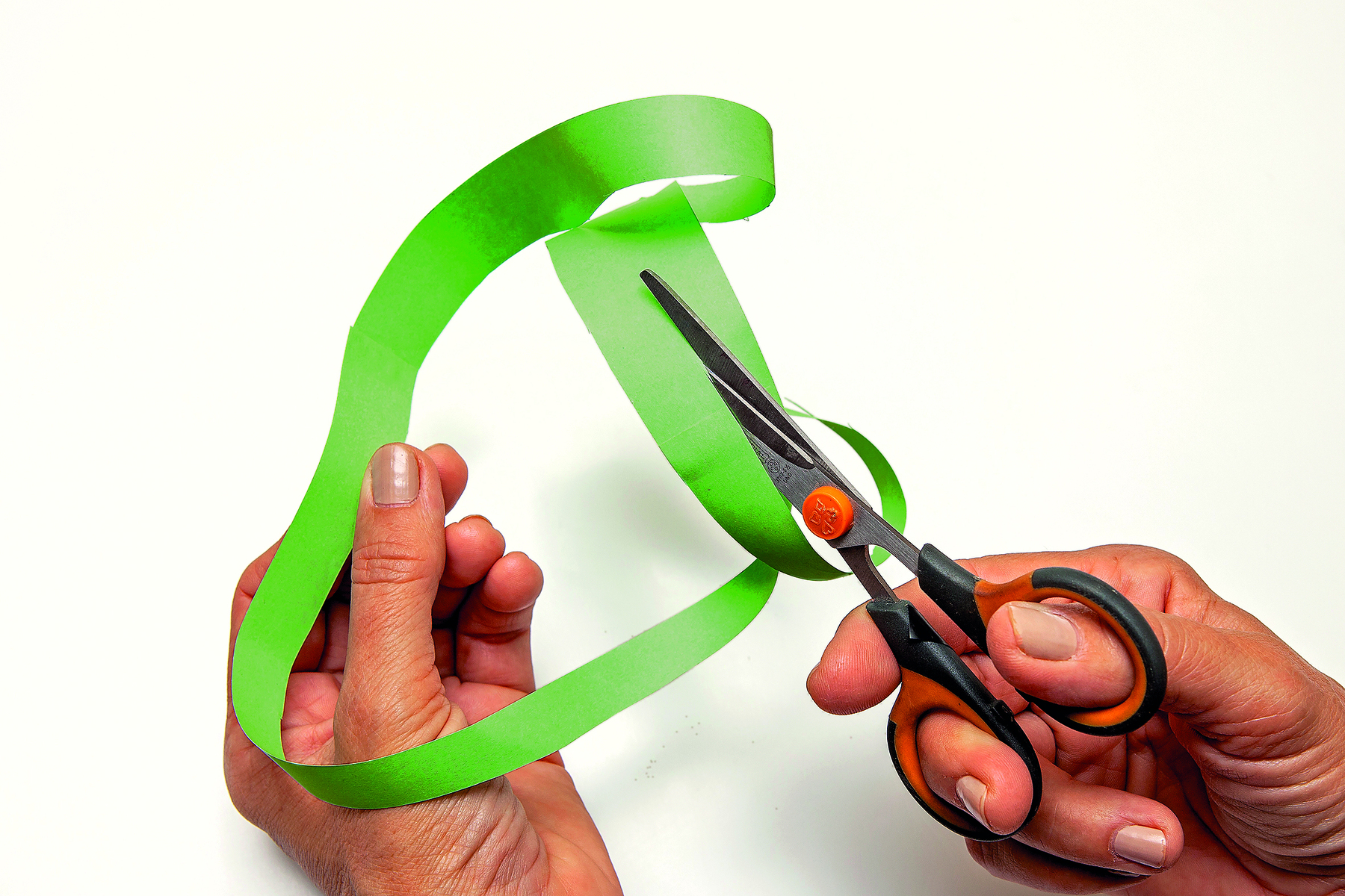 Fotografia. Detalhe de mãos cortando a tira de papel verde em tiras mais finas.
