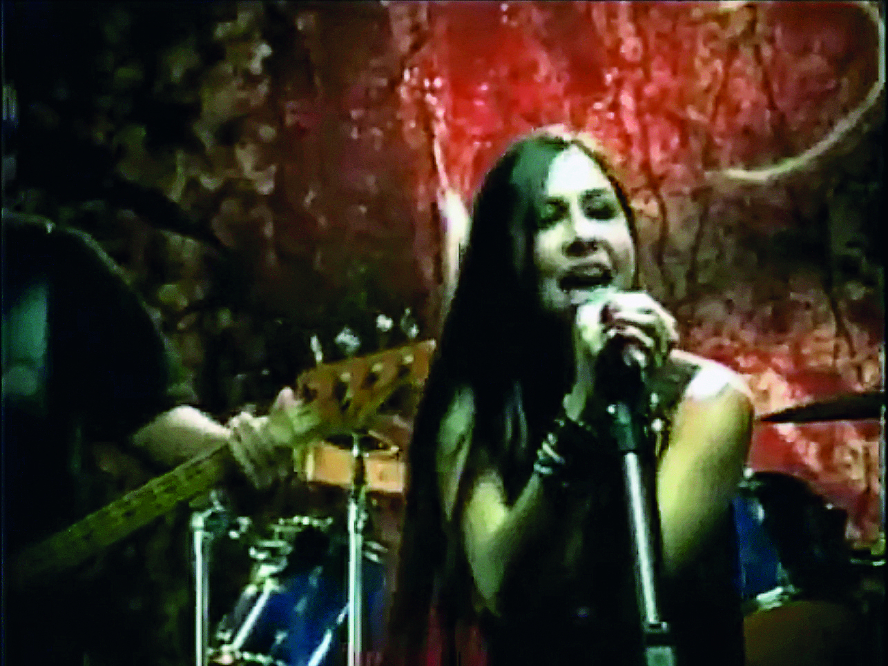 Fotografia. Mulher de cabelo longo castanho, vestida com camiseta regata preta, canta ao microfone. Ao fundo, cenário do palco em tons avermelhados.