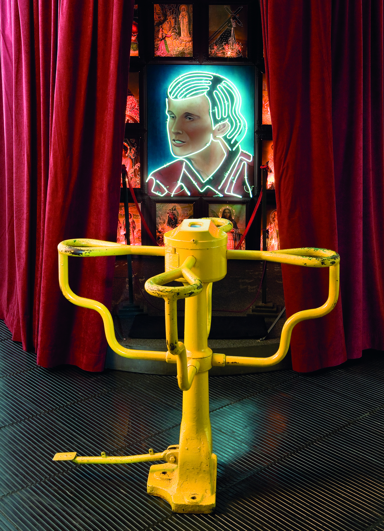 Fotografia. Obra de arte feita com uma catraca amarela. Acima da catraca, uma tela com o desenho de rosto masculino com contornos luminosos feito com lâmpadas neon. Ao redor da tela, cortinas vermelhas.
